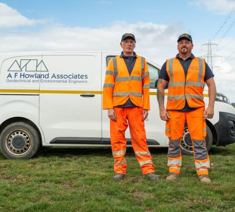 Two A F Howland Associates engineers in high-visibility PPE standing beside a branded geotechnical engineering van on a rural project site.